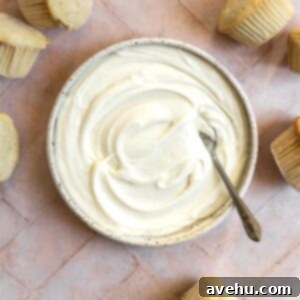 Cream cheese frosting in a bowl next to unfrosted cupcakes on a pink tiled surface.