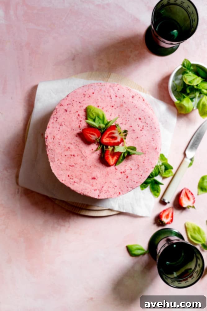 Aromatic Strawberry Basil Cake with Strawberry Buttercream 14 A beautifully frosted pink strawberry cake, topped with fresh strawberries and basil, sits on parchment paper against a soft pink background.