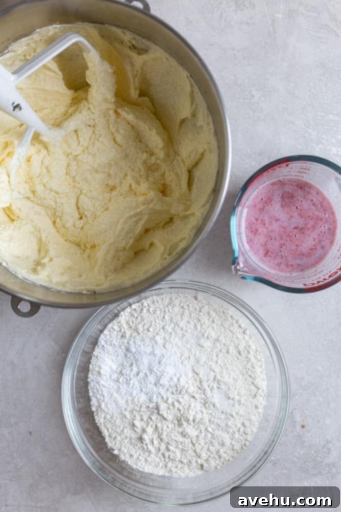 Aromatic Strawberry Basil Cake with Strawberry Buttercream 5 A mixing bowl filled with creamy, pale yellow cake batter, flanked by a bowl of dry flour mixture and a measuring cup containing a red strawberry-milk blend, all on a gray surface.