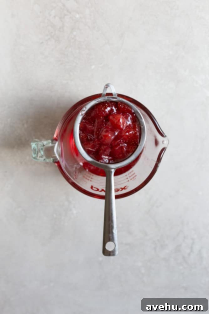 Aromatic Strawberry Basil Cake with Strawberry Buttercream 8 A fine-mesh strainer sits atop a clear measuring cup, filled with cooked strawberries, allowing the rich strawberry simple syrup to collect below.