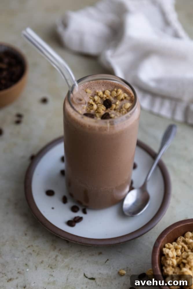 Energizing 3-Minute Greek Yogurt Coffee Smoothie 2 A coffee smoothie in a glass cup with glass straw on a plate next to a spoon, topped with granola.