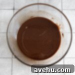 Handcrafted Fudgy Chocolate Brownie Masterpiece 4 Close-up of melted chocolate and butter in a clear glass bowl, glistening on a light-colored tile surface, ready to be mixed into the brownie cake batter.