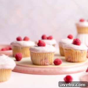 Seriously Moist Homemade Raspberry Cupcakes 2 Moist raspberry cupcakes on an overturned pink plate, garnished with fresh raspberries and a swirl of tangy cream cheese frosting.