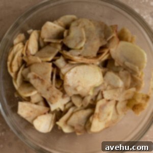 Thinly sliced Honeycrisp apples, generously coated with a sweet and aromatic cinnamon-brown sugar mixture, gathered in a large clear glass mixing bowl, ready for baking.