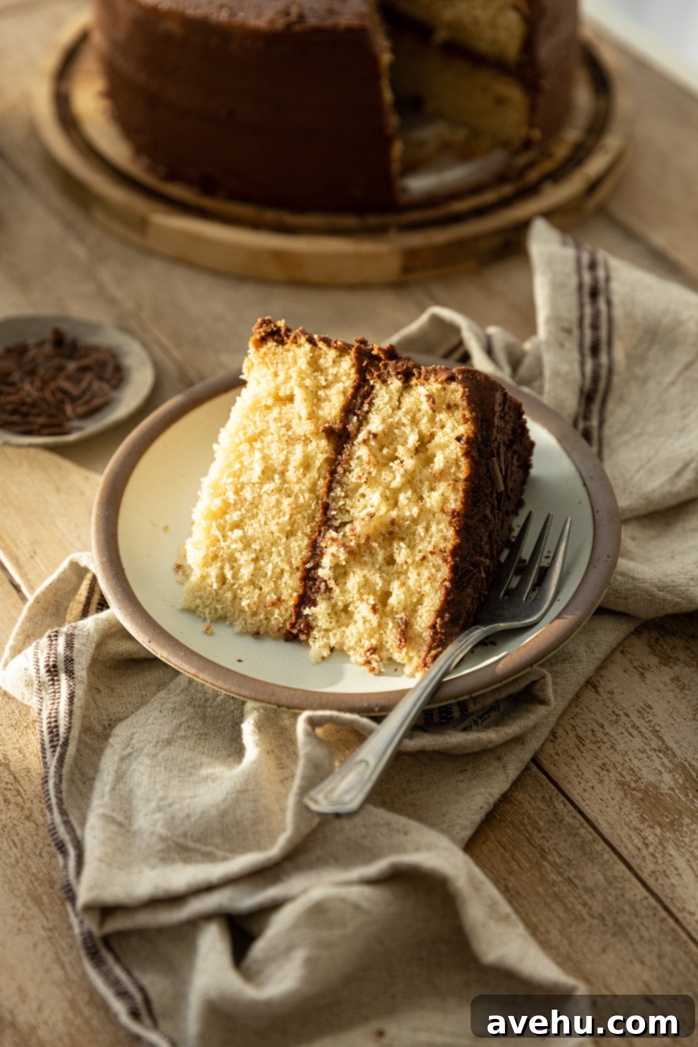 A slice of yellow cake with chocolate frosting on a rustic plate and linen napkin.