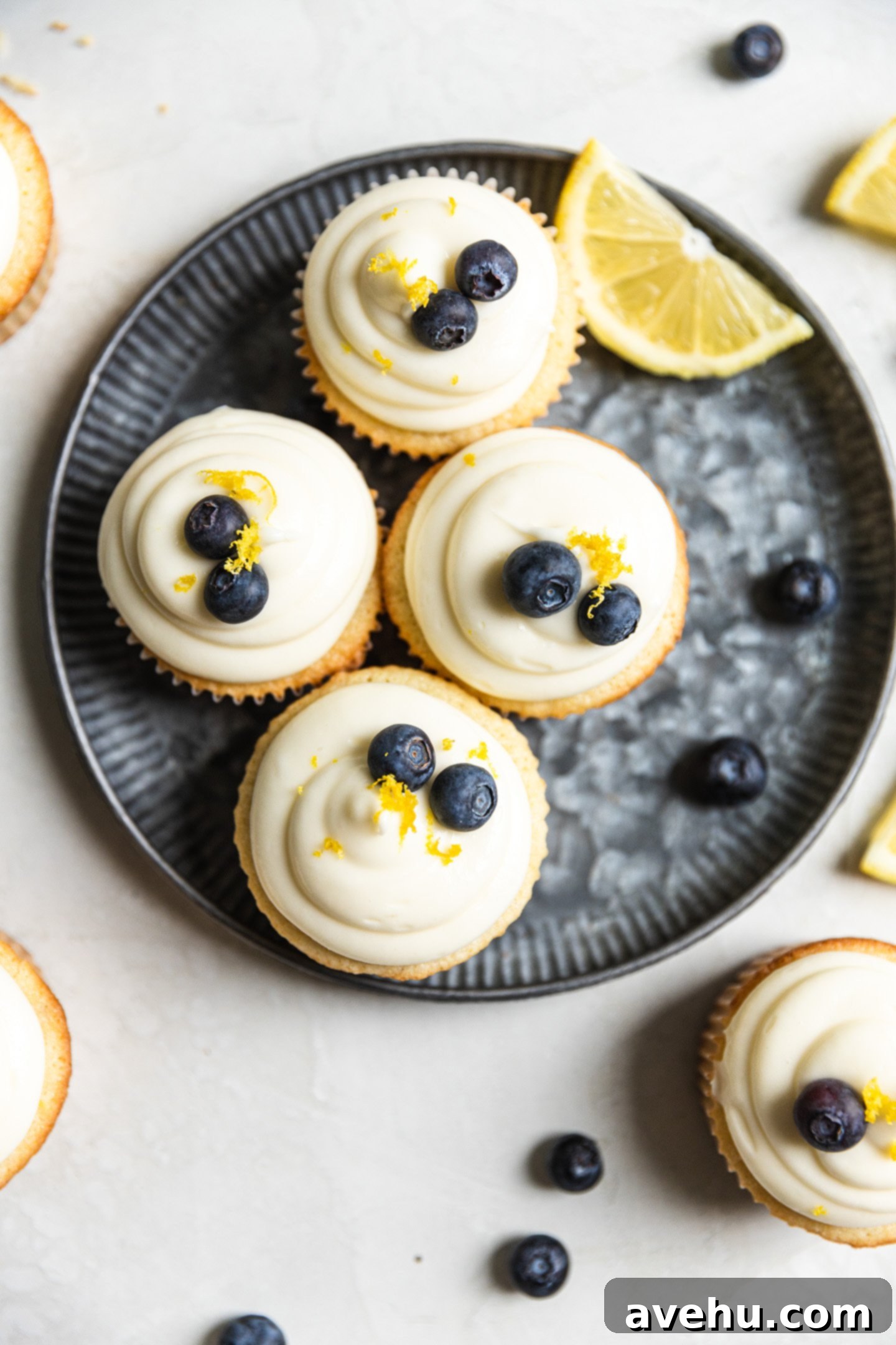 Bright & Zesty Lemon Blueberry Cupcakes 2 Four beautifully frosted lemon blueberry cupcakes arranged on a blue-tinted plate, accompanied by fresh lemon slices and a scattering of vibrant blueberries, ready for serving.