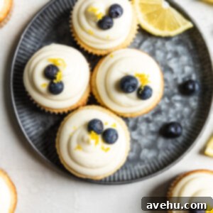 Bright & Zesty Lemon Blueberry Cupcakes 11 Four lemon blueberry cupcakes on a blue tinted plate with a slice of lemon and blueberries next to them.