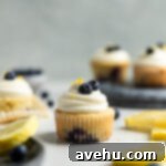 Bright & Zesty Lemon Blueberry Cupcakes 19 A lemon blueberry cupcake frosted with white frosting on a table next to other frosted cupcakes and slices of lemons.