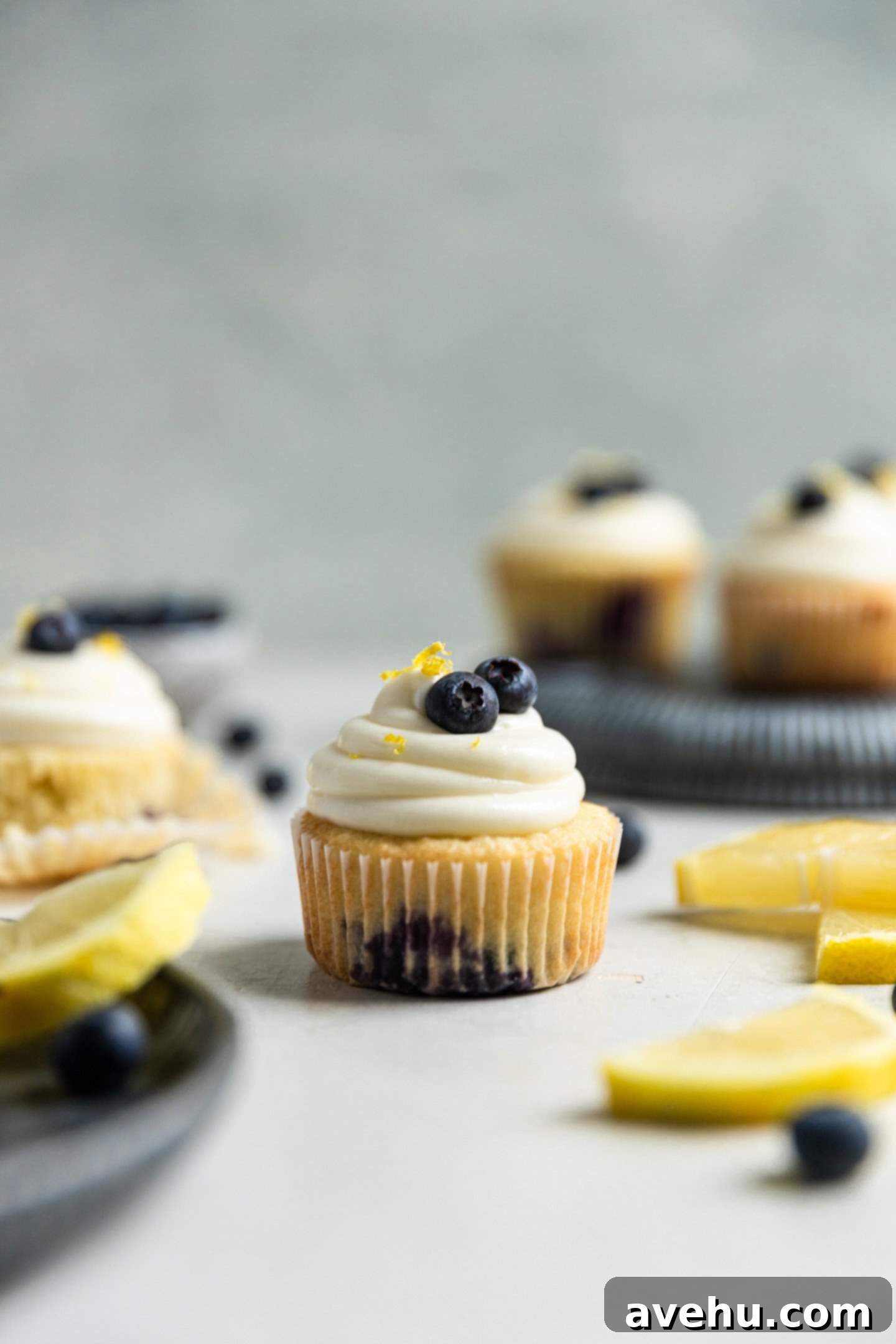 Bright & Zesty Lemon Blueberry Cupcakes 10 A single beautifully frosted lemon blueberry cupcake, garnished with fresh blueberries and lemon zest, stands invitingly on a table alongside other frosted cupcakes and bright lemon slices.