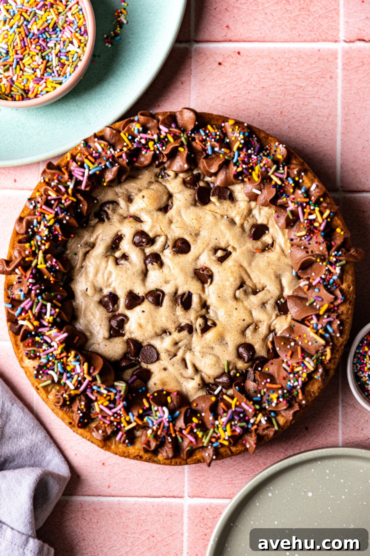 Effortless Homemade Chocolate Chip Cookie Cake 2 A chocolate chip cookie cake decorated with chocolate frosting and rainbow sprinkles on a pink tile surface.