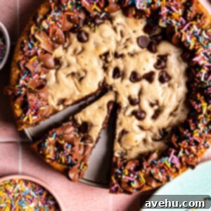 Effortless Homemade Chocolate Chip Cookie Cake 3 A chocolate chip cookie cake with chocolate frosting and sprinkles with a slice cut out on a pink background.