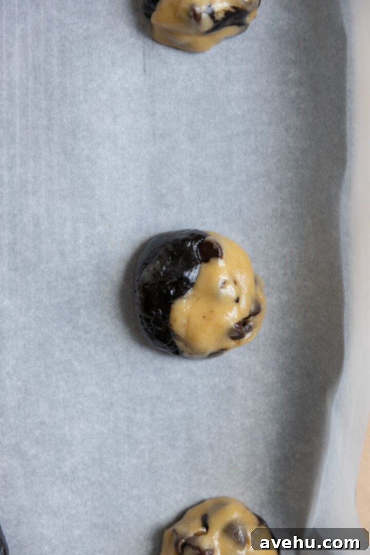 A swirl of brownie and chocolate chip cookie dough formed into a single, perfectly round ball, nestled on a parchment-lined cookie sheet, ready for baking.