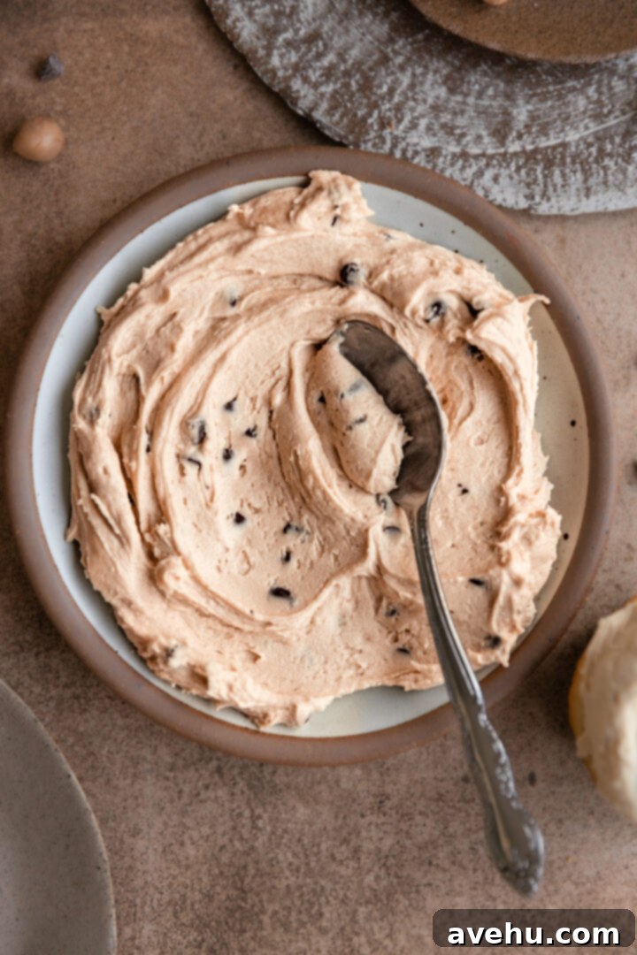 Silky Smooth Cookie Dough Frosting 2 A plate of creamy, perfectly spreadable cookie dough frosting, adorned with mini chocolate chips, with a spoon having taken a scoop from it.
