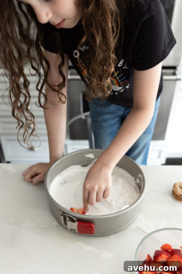 Chilled Strawberry Cloud Cake 8 A child carefully placing vibrant, sliced strawberries on a layer of whipped cream within a springform pan, constructing the icebox cake.
