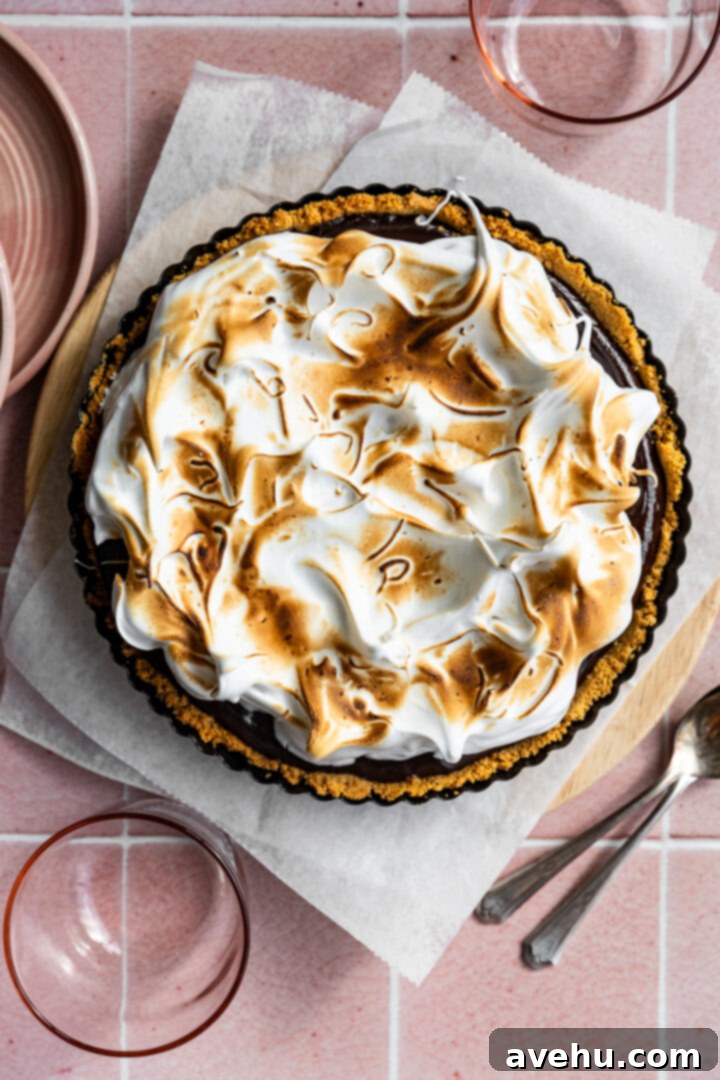 Foolproof S'mores Pie with Decadent Chocolate 2 A s'mores pie with torched meringue set on a pink tiled backdrop with plates, spoons, and cups around.