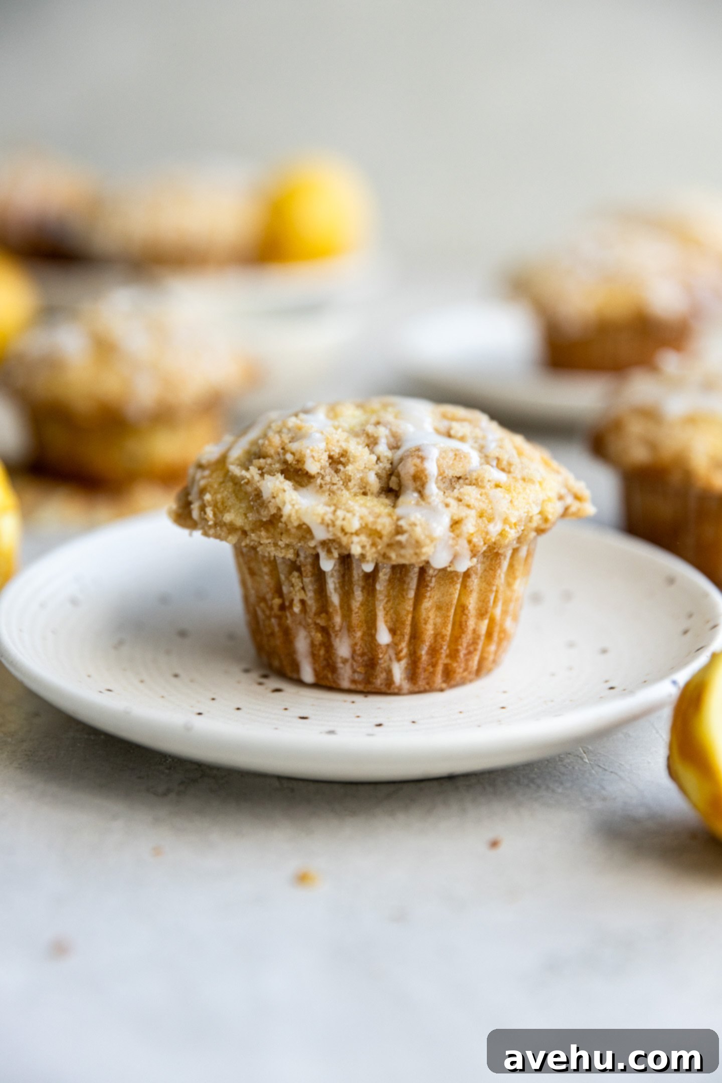Gourmet Triple Lemon Muffins 2 A perfectly baked, domed lemon muffin on a white spotted plate, with more bakery-style lemon muffins blurred in the background, showcasing their golden tops and glistening glaze.