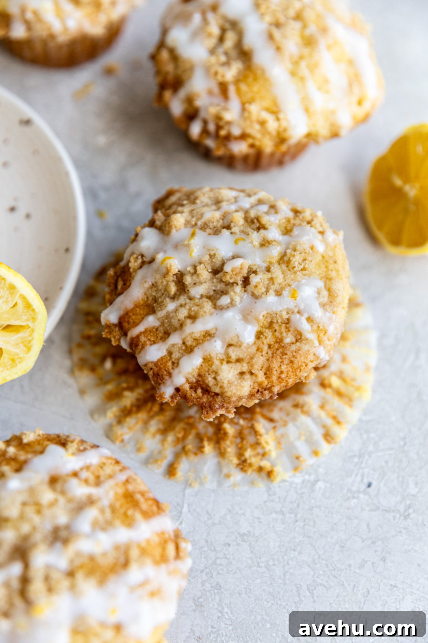 Gourmet Triple Lemon Muffins 12 A glistening lemon streusel muffin with its paper liner partially peeled, resting on a wire rack with other glazed muffins in the background, showing its tender interior.