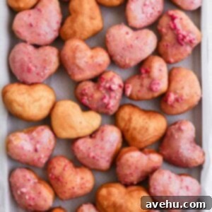 Sweetheart Strawberry Vegan Donuts 3 A vibrant assortment of strawberry heart-shaped donuts, some elegantly glazed and others simply fried, displayed on a baking sheet.