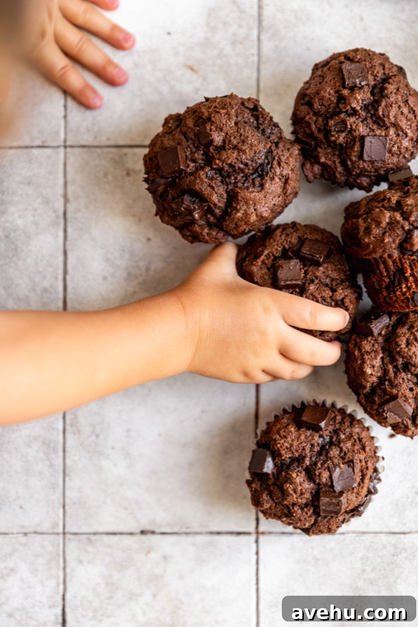 Ultimate Bakery Triple Chocolate Muffins 9 A childs hand reaching for a muffin on a tiled surface.