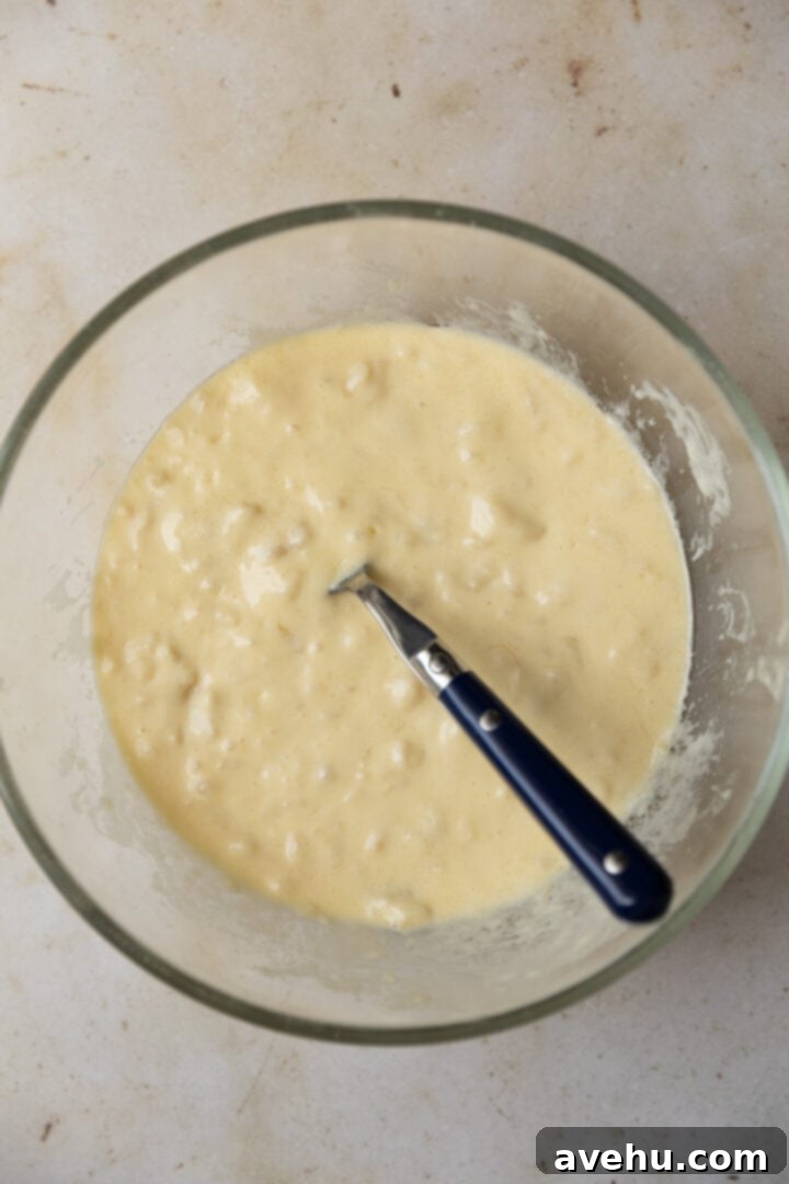 Perfectly Moist Bakery-Style Banana Muffins 5 Wet ingredients for banana muffins (melted butter, mashed bananas, oil, yogurt, eggs, vanilla) in a large bowl, ready to be whisked.