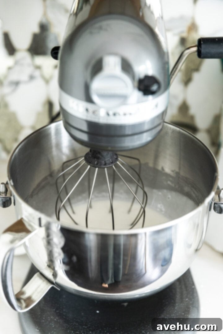 Decadent Cocoa Swiss Meringue Buttercream 5 A stand mixer bowl with stiff, glossy meringue being whipped by a whisk attachment.