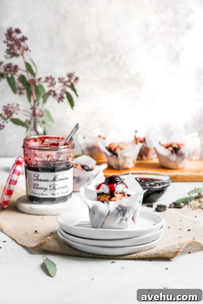 Nutty Butter Dark Cherry Chocolate Muffins 2 A perfectly baked cherry muffin sitting on a stack of pristine white plates, with an open jar of Bonne Maman Cherry Preserves subtly blurred in the background.