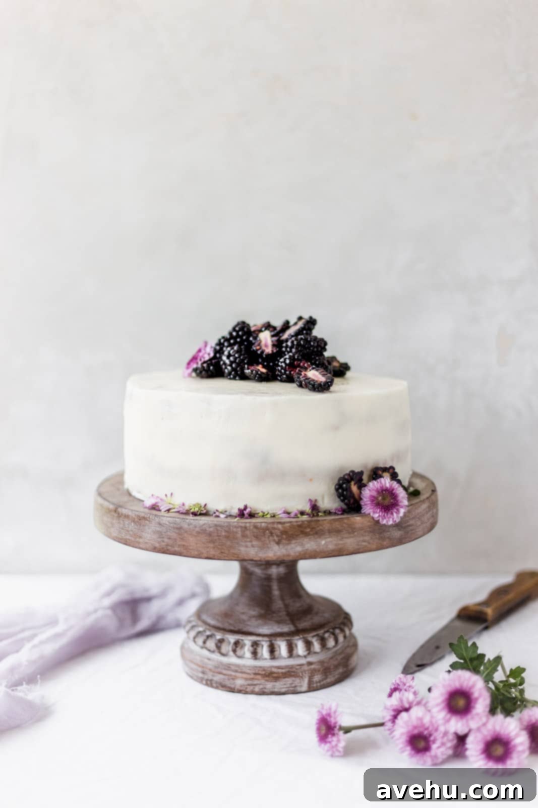 Effortless and Perfectly Moist Blackberry Cake 2 White cake on a brown cake stand with purple flowers and blackberries, showcasing a beautiful homemade blackberry cake.