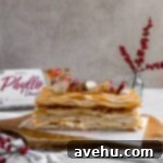 A Napoleon dessert made with phyllo dough on a wooden cutting board topped with dried apples and red flower berries in the background.
