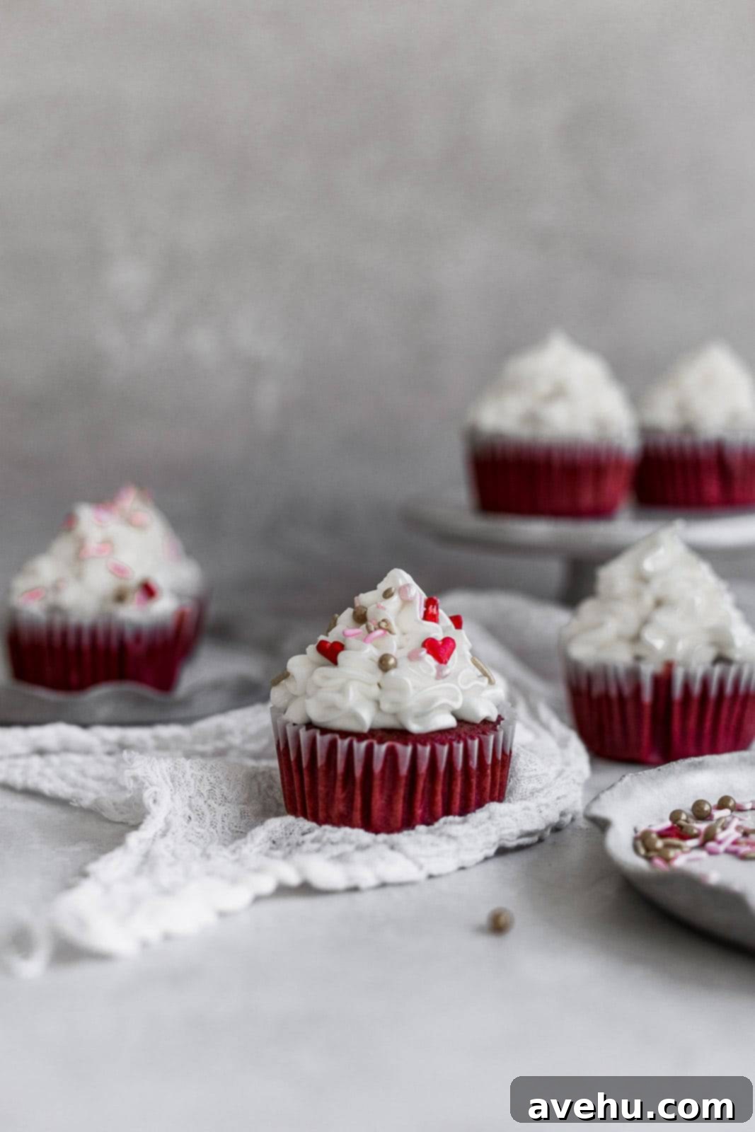 Scarlet Velvet Cheesecake Delights 2 A vibrant red velvet cupcake topped with fluffy white cream cheese frosting and festive sprinkles, resting on white gauze with other cupcakes blurred in the background on a gray surface, perfect for Valentine's Day.
