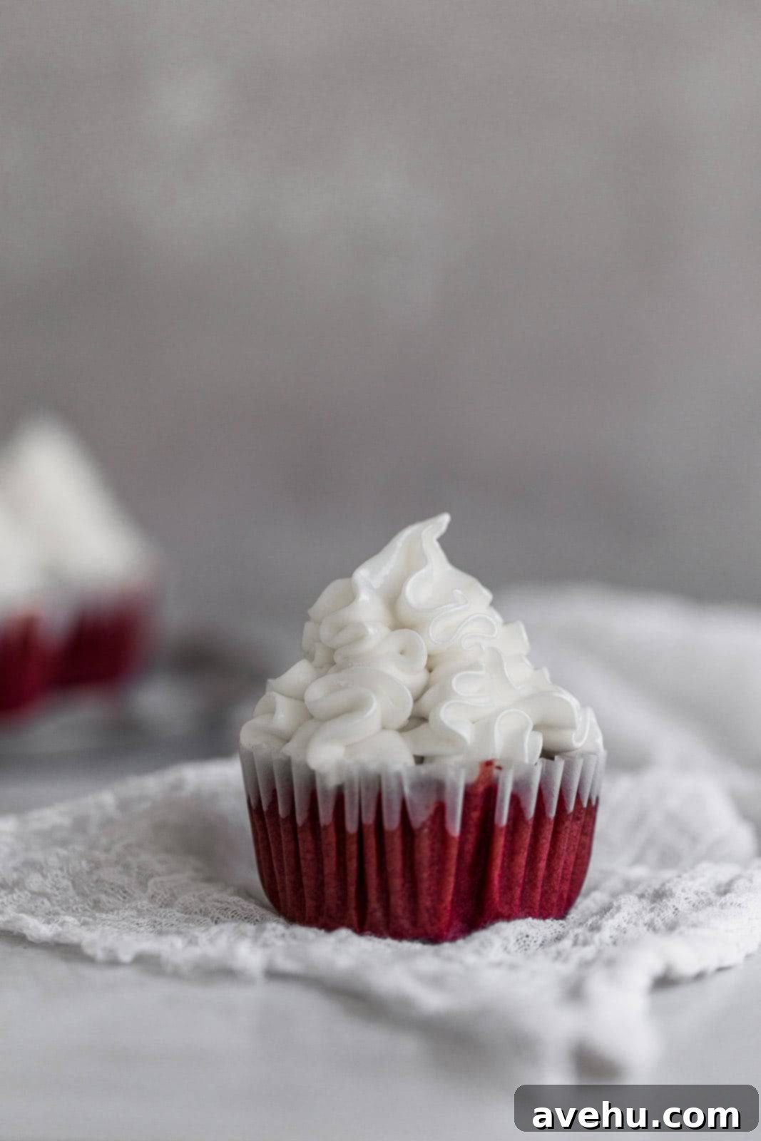 Scarlet Velvet Cheesecake Delights 6 A single red velvet cupcake with perfectly piped cream cheese frosting on top, resting on white cheesecloth against a soft gray background, showcasing its inviting appearance.
