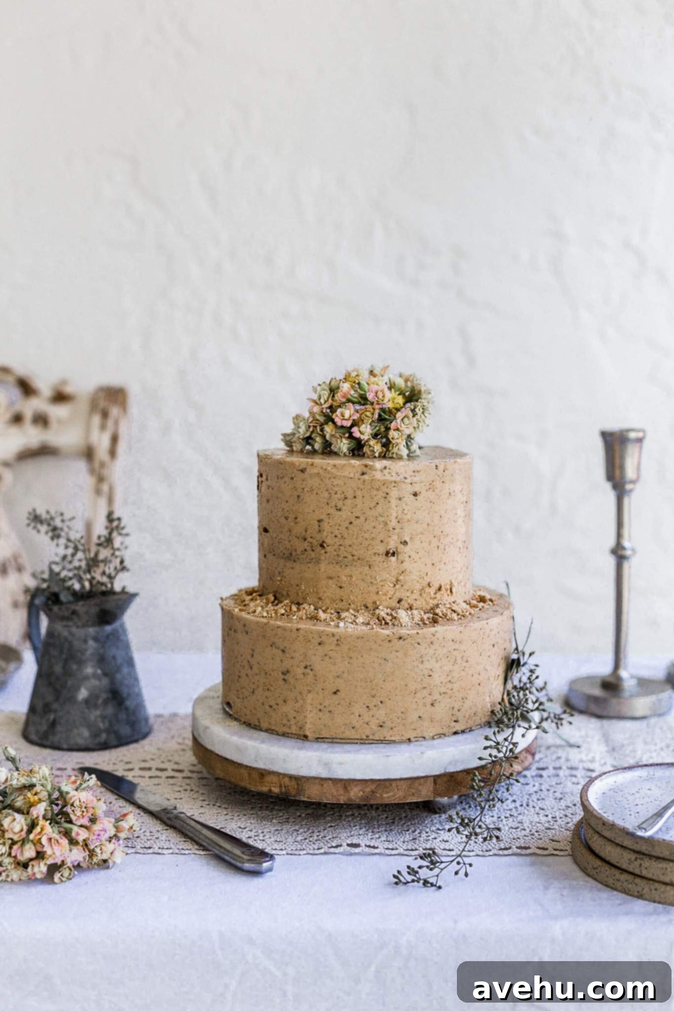 Mastering Two-Tier Cakes: Baking and Stacking Techniques 2 A two tier cookie butter cake on a marble and wood cake plate sitting on a white eyelet tablecloth decorated with pink flowers on top.