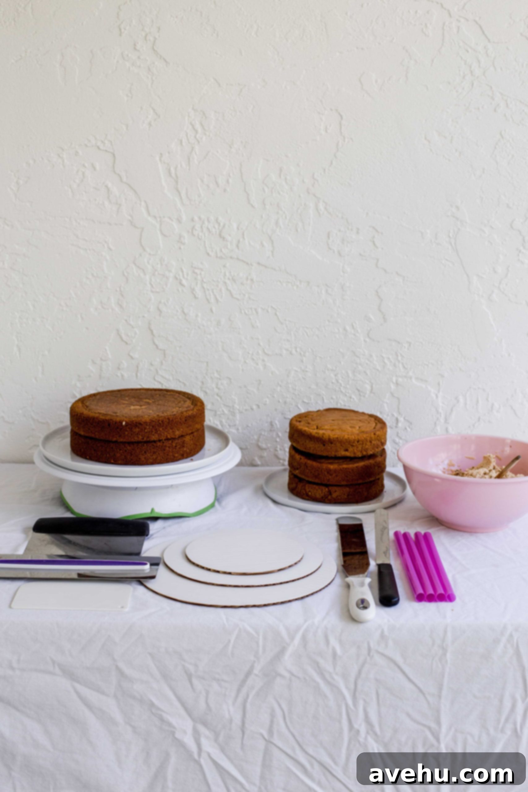 Mastering Two-Tier Cakes: Baking and Stacking Techniques 6 A table full of things needed to stack a cake including 2 cakes, cake boards, bench scrapers, buttercream in a pink bowl, bubble straws, a long serrated knife, and an offset spatula