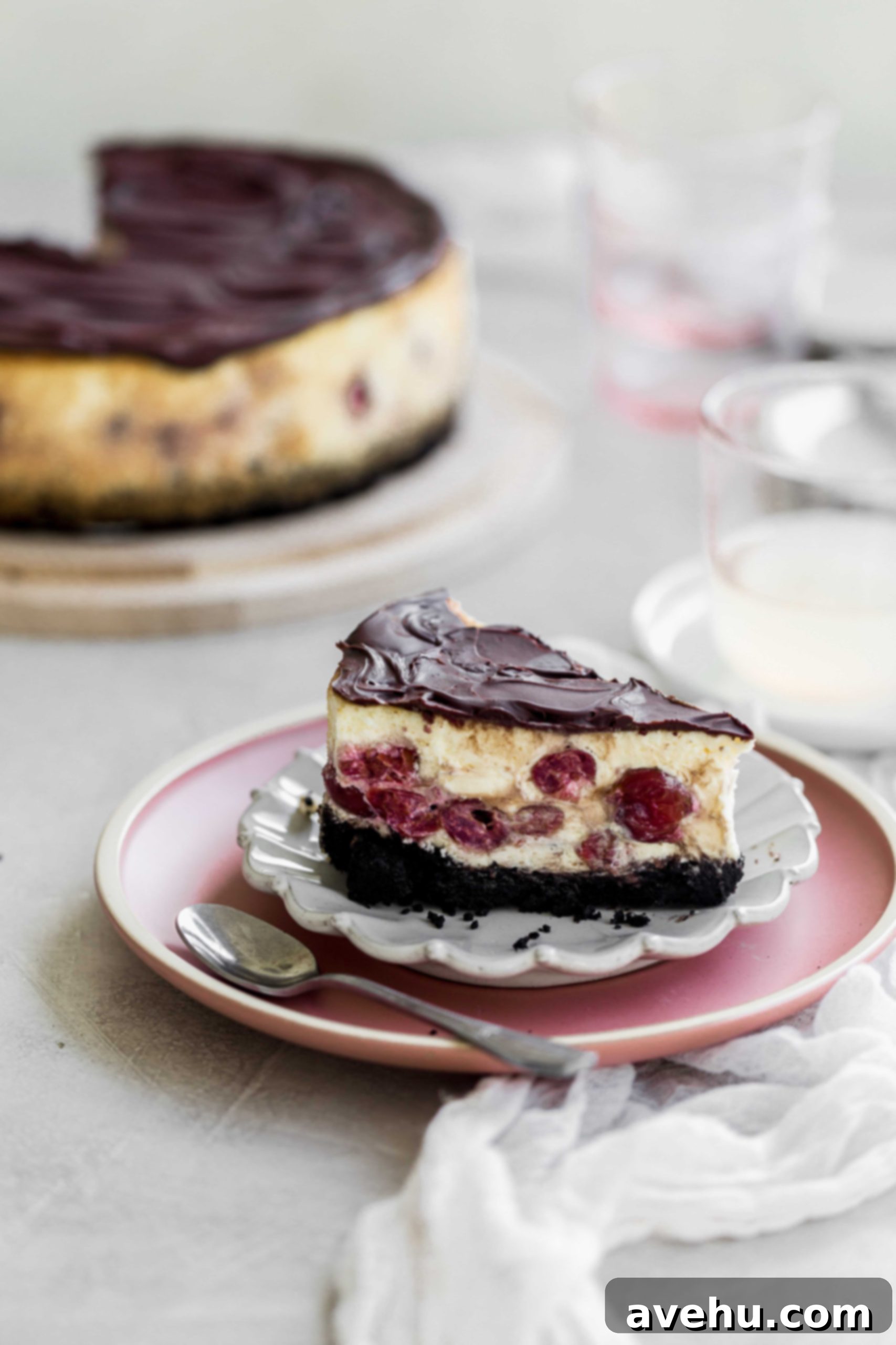Decadent Cherry Chocolate Cheesecake 2 A slice of chocolate cheesecake with cherries in the middle sitting on two plates. The larger one is pink the smaller one is gray. There is a spoon on the pink plate.