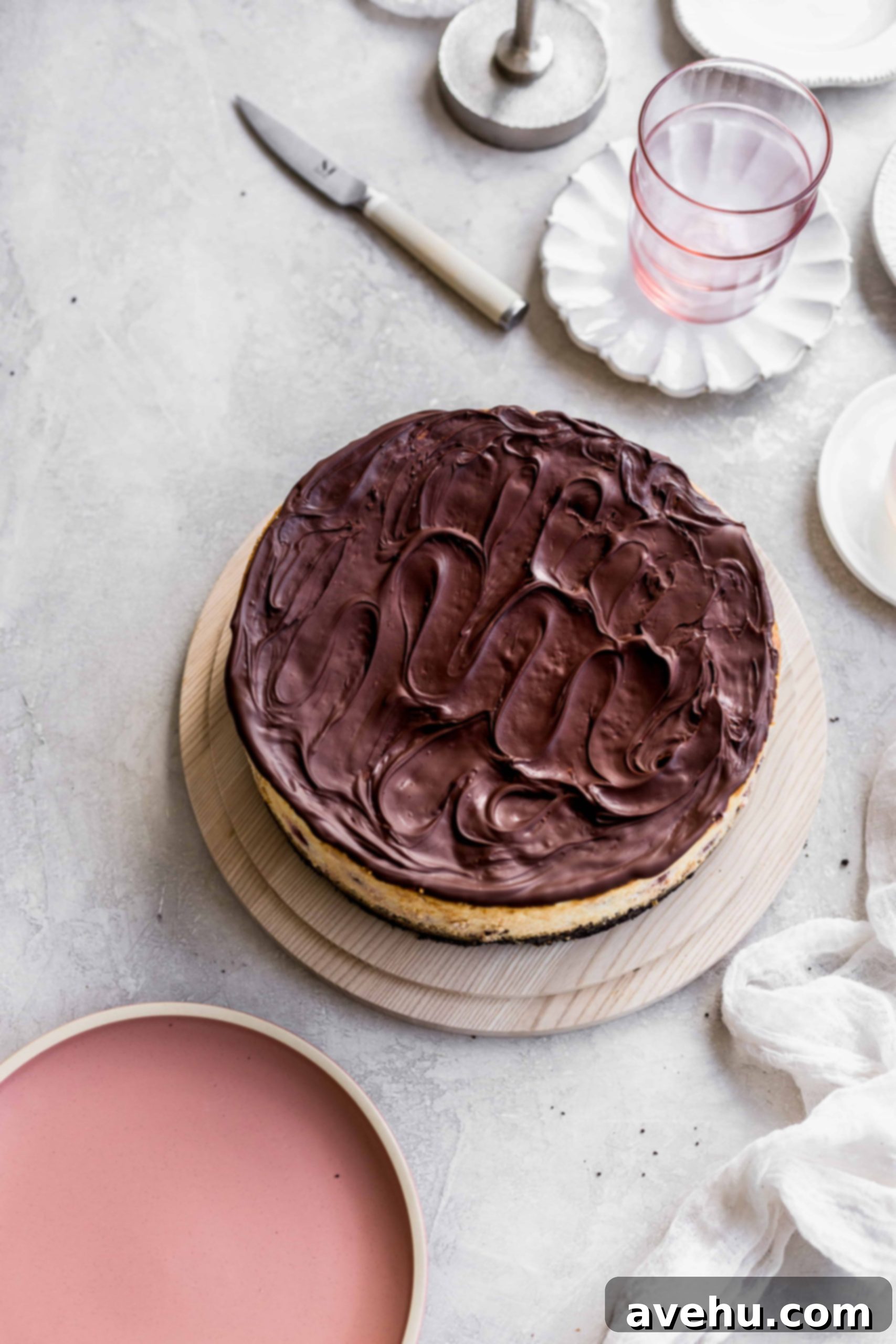 Decadent Cherry Chocolate Cheesecake 3 Chocolate swirled all over the top os a cheesecake on a wood cake plate on a gray background with pink plates around it.