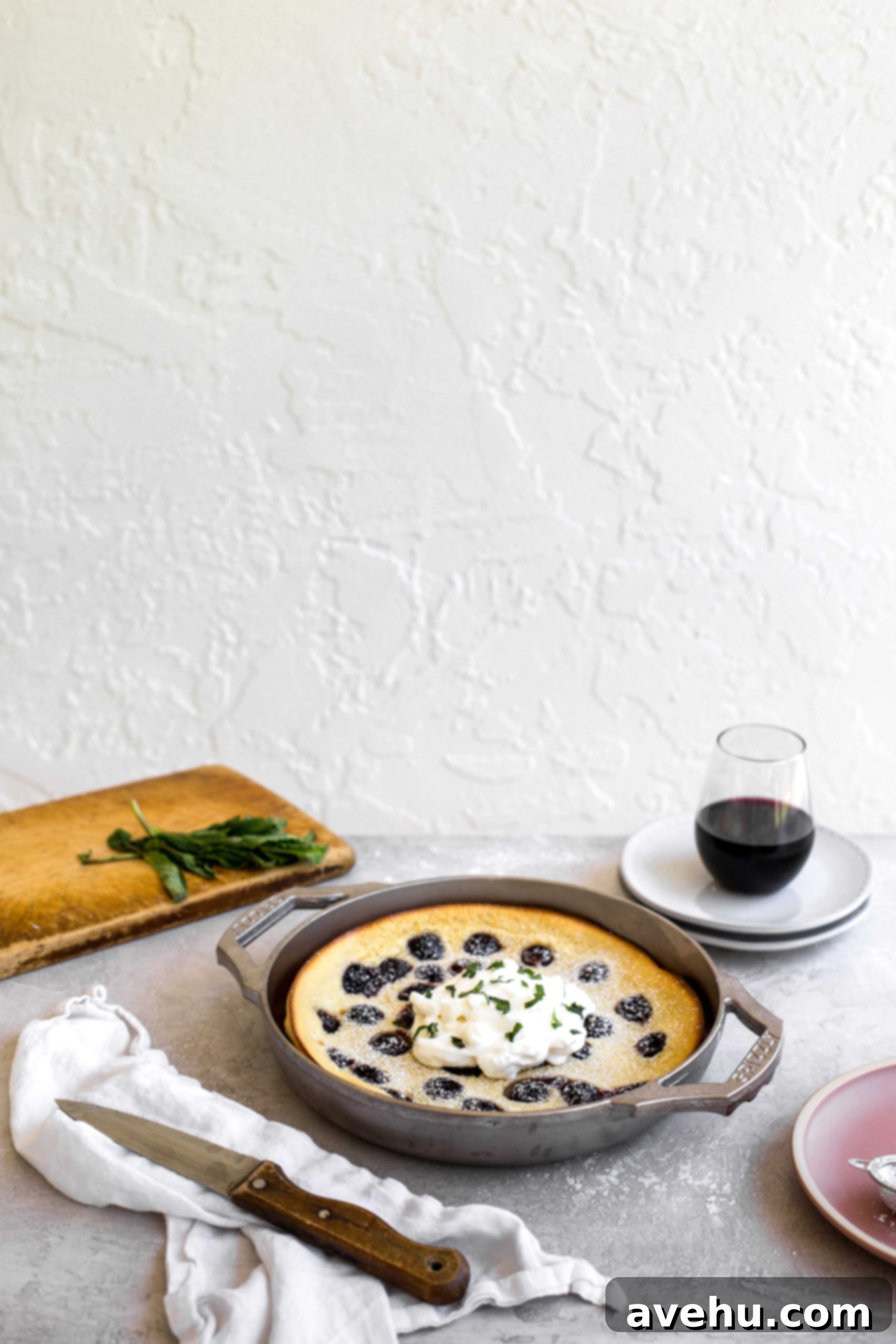 Bourbon Cherry Clafoutis 2 The side view of a cherry clafoutis made in a GRIZZLY nickel plated cast iron pan set on a gray table with a cutting board in the background and a cup of wine on some plates.