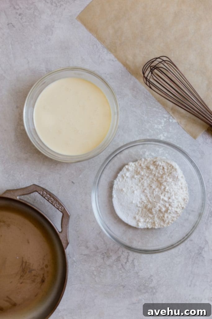 Bourbon Cherry Clafoutis 5 Flour in a glass bowl next to a yellow batter in another glass bowl with a cast iron pan next to it and a white whisk on the side on top of a grey surface.