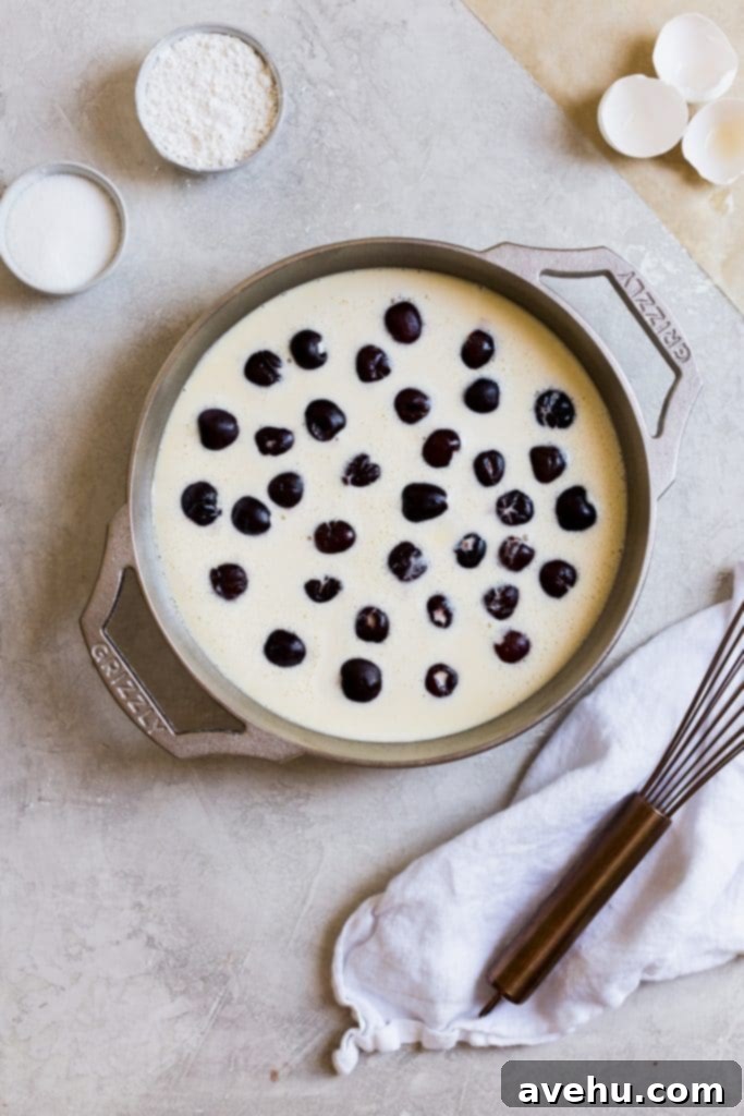 Bourbon Cherry Clafoutis 6 A GRIZZLY cast iron casserole dish filled with a yellow batter and scattered with frozen cherries on a gray background.