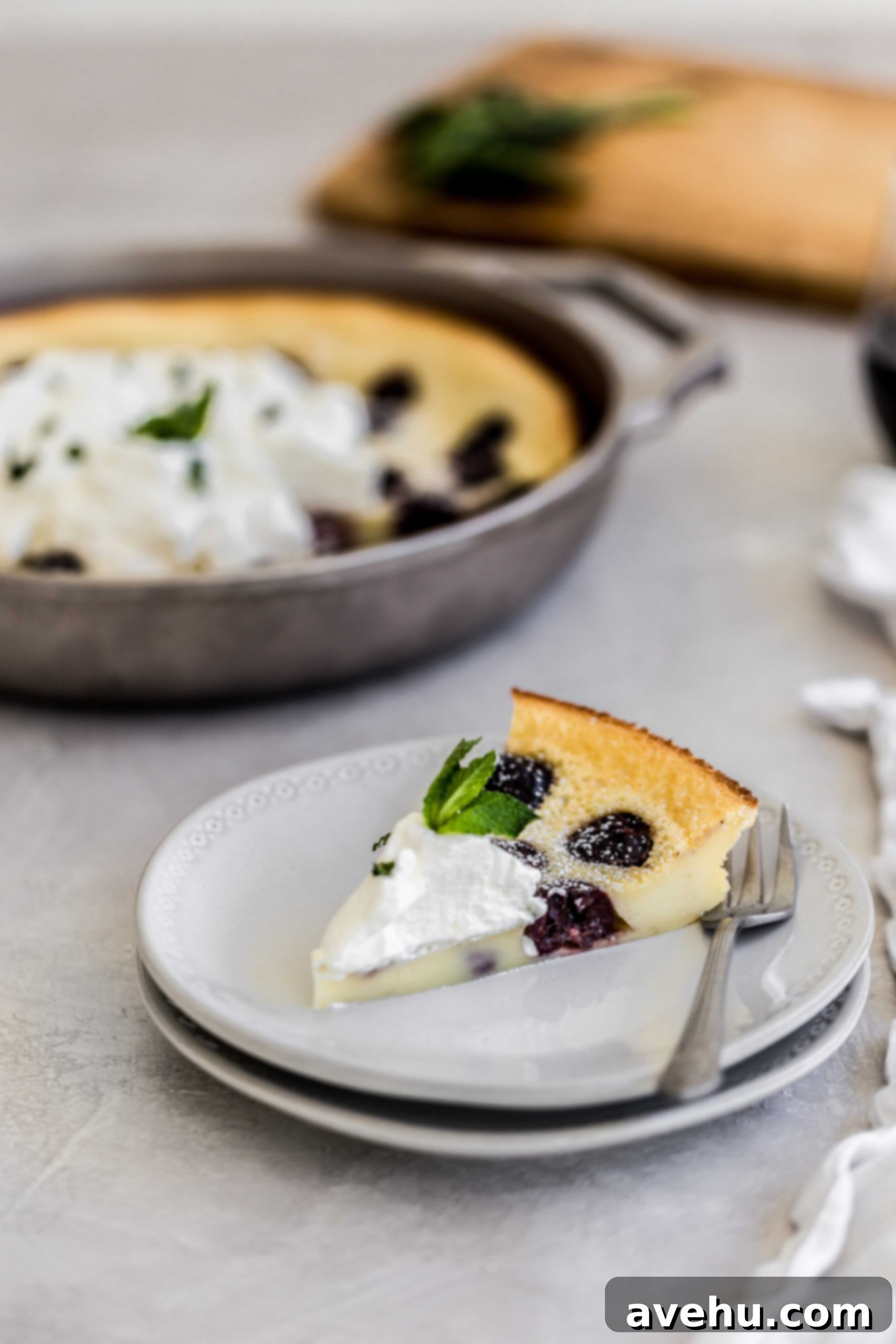 Bourbon Cherry Clafoutis 9 a slice of cherry clafoutis on a pair of grey stacked plates with the ;leftovers in the pan in the background of a grey surface.