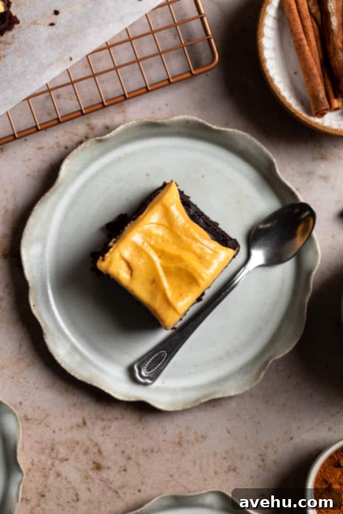 Decadent Chocolate Pumpkin Sheet Cake 6 A perfectly sliced piece of chocolate pumpkin sheet cake, topped generously with pumpkin cream cheese frosting, rests on a gray plate with a spoon beside it.