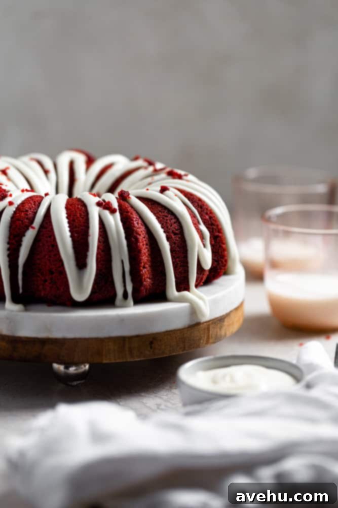 Decadent Fluffy and Moist Red Velvet Bundt Cake 4 A thick and creamy cream cheese frosting beautifully drizzled over a freshly baked red velvet bundt cake, with soft pink cups subtly blurred in the background, creating an inviting scene.