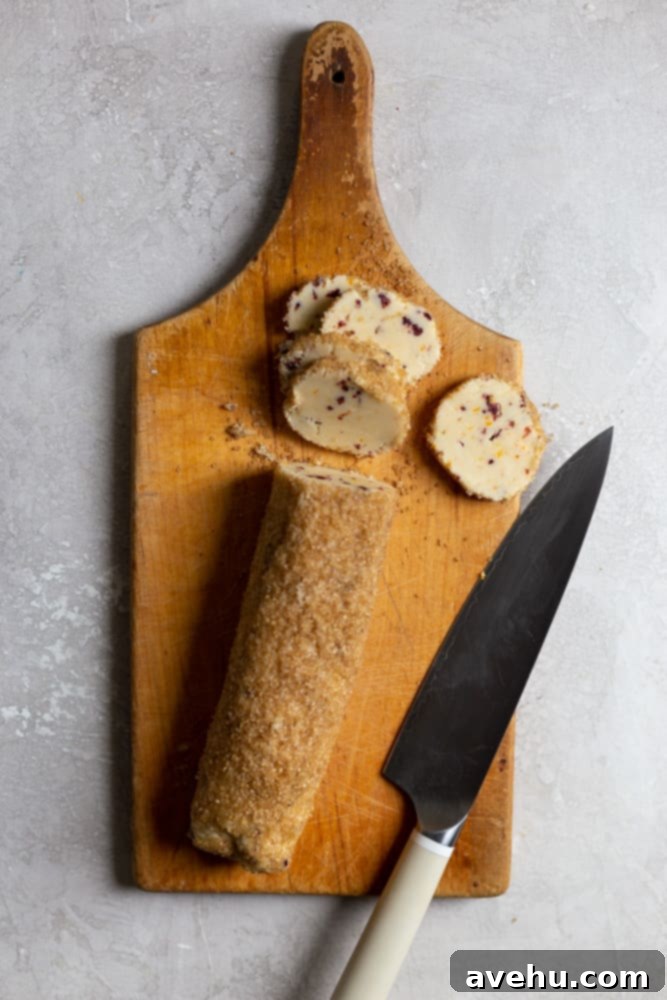 Cloudlike Cranberry Shortbread Delights 7 A well-chilled, sugar-coated log of cranberry orange shortbread dough resting on a wooden cutting board, poised for precise slicing.