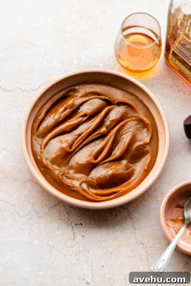 Foolproof Bourbon Caramel Sauce Visual Guide 2 A beautiful pink bowl filled with luscious homemade bourbon caramel sauce, gracefully positioned on a white marble surface. A glass containing bourbon sits subtly in the background, hinting at the key ingredient within the rich, amber sauce.