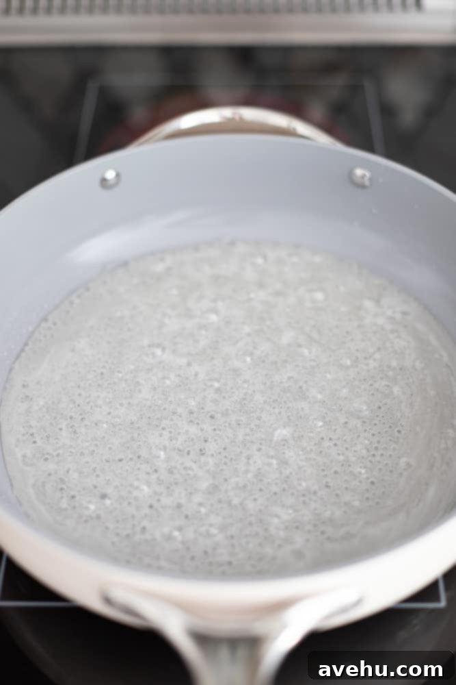 Foolproof Bourbon Caramel Sauce Visual Guide 5 Small, gentle bubbles starting to form in the sugar and water mixture at the bottom of a light gray pot, indicating the beginning of the heating process.